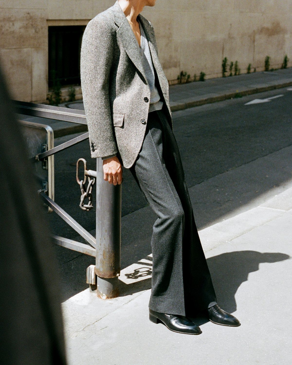 A person wearing a grey textured blazer and dark trousers leaning against a metal railing.