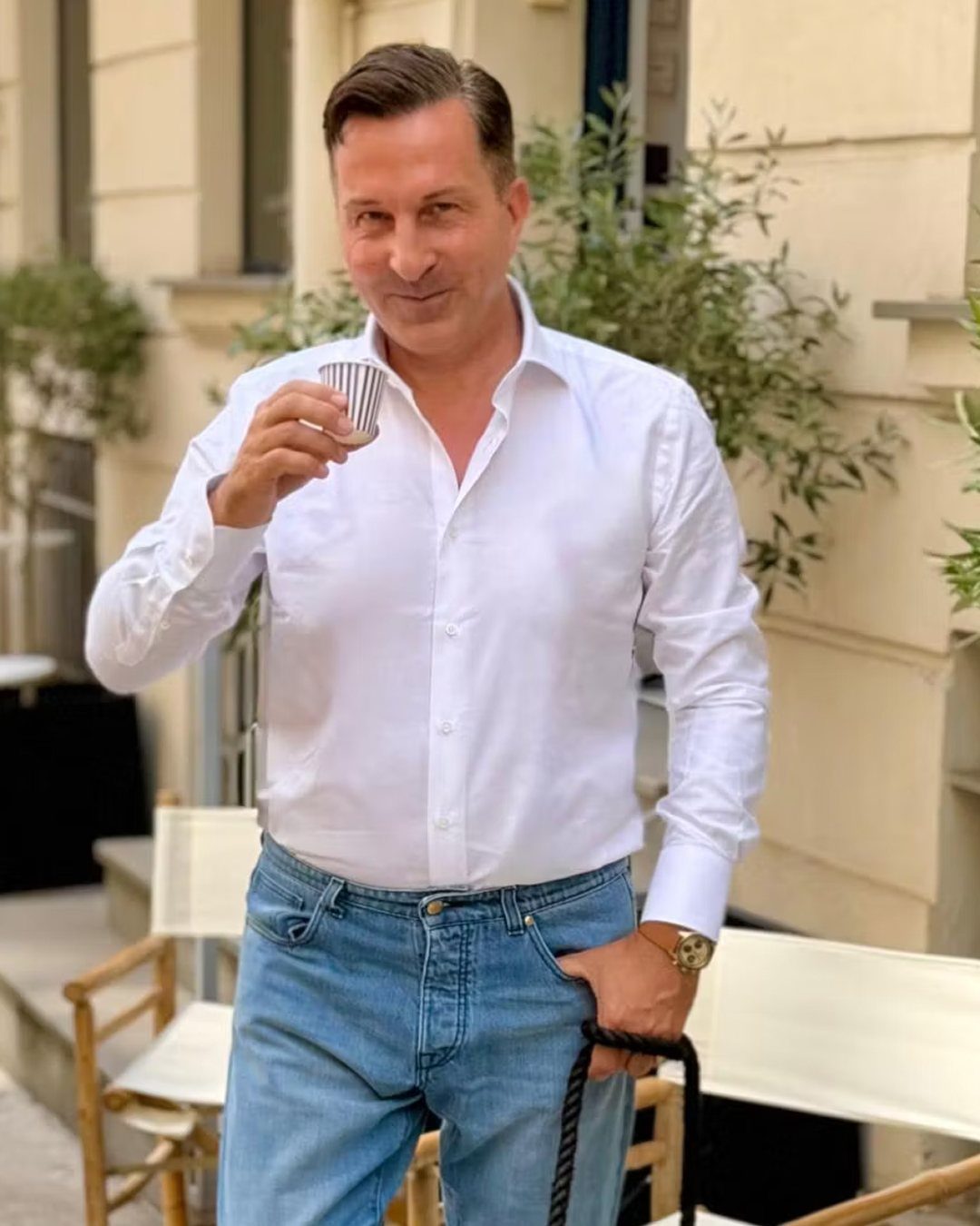 A man in a white button-down shirt and blue jeans holding a small espresso cup.