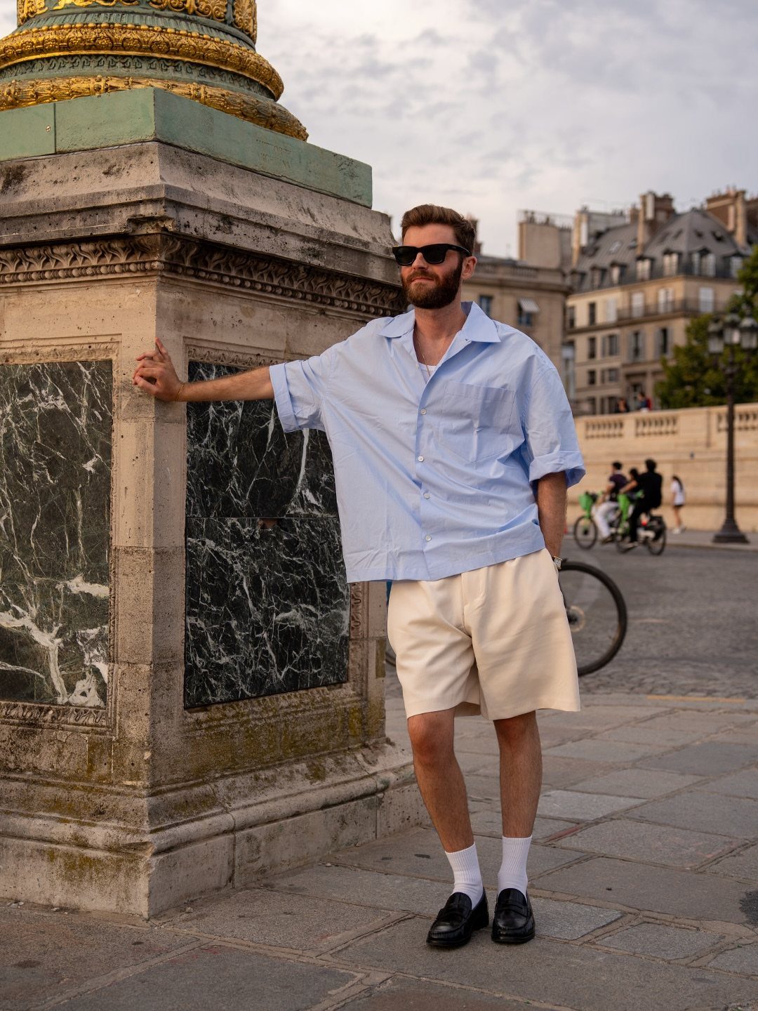 A man in a light blue button-down shirt and cream shorts leaning against a stone monument.