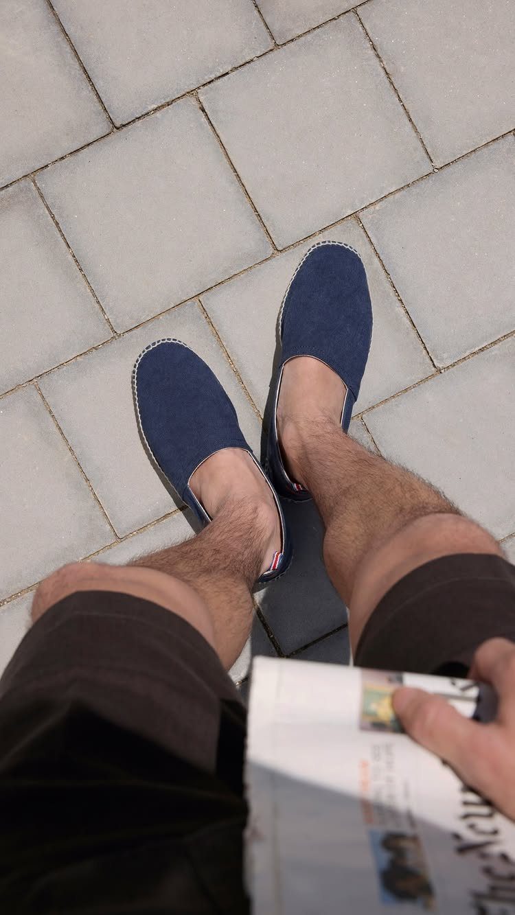 Navy blue slip-on espadrilles worn by a person standing on grey tiles holding a newspaper.