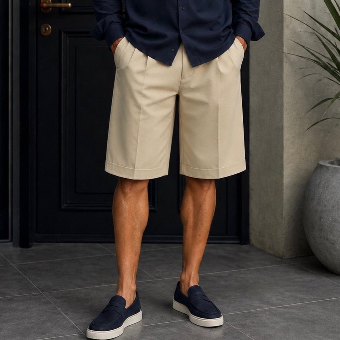 Man wearing beige pleated tailored shorts, a navy shirt, and navy loafers standing outdoors.