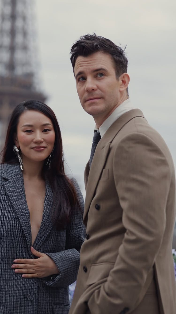 A man in a tan blazer and a woman in a grey patterned blazer posing outdoors.