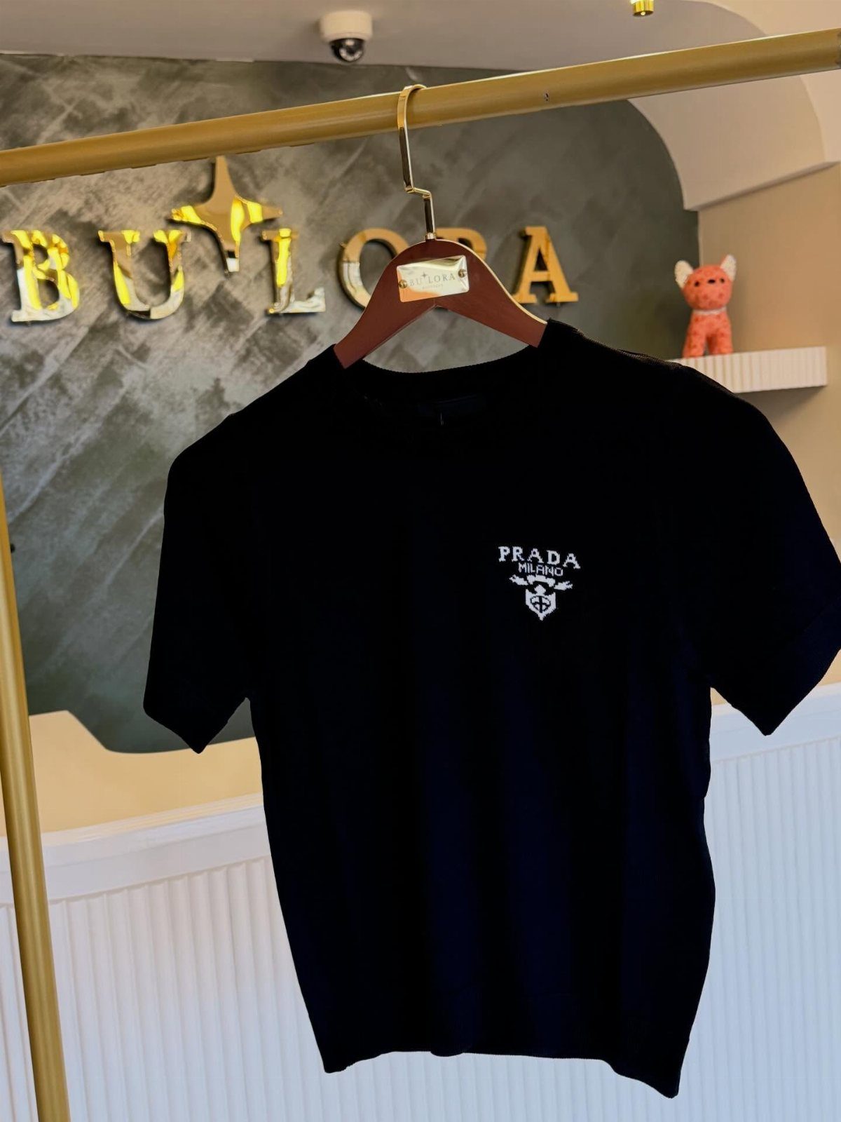 Black Prada short-sleeve t-shirt with white embroidered logo hanging on a gold clothing rack.