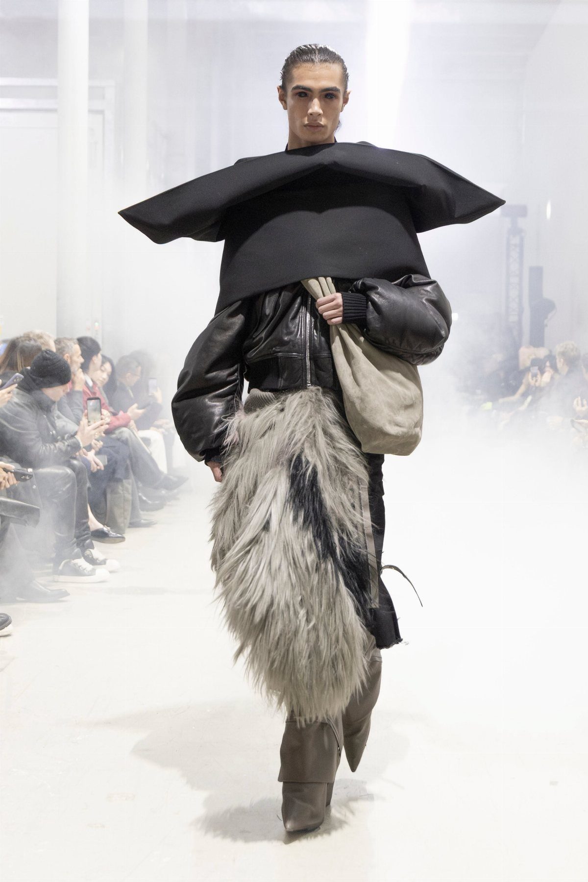 Model walking a runway in avant-garde black structured top and long grey fur skirt.