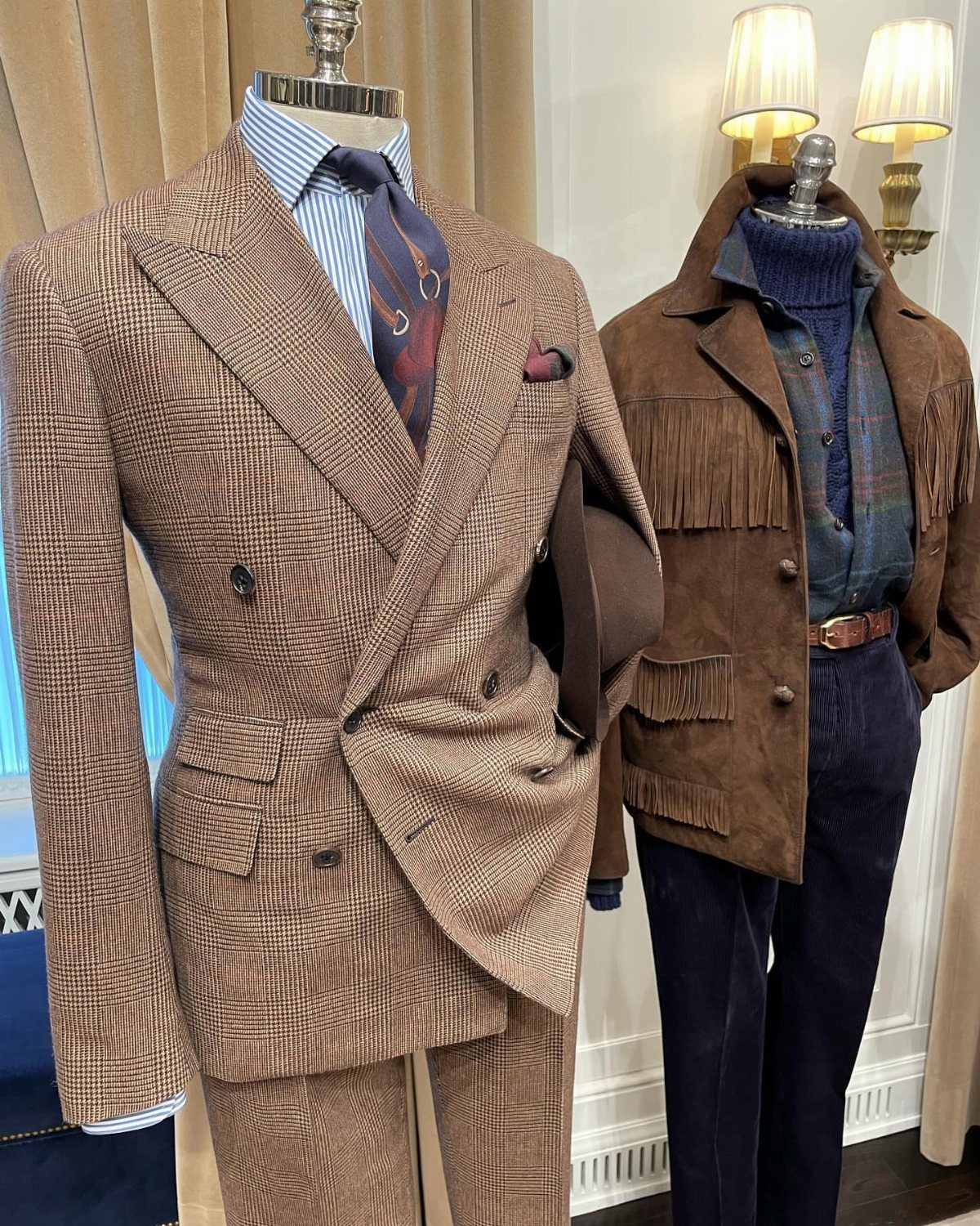 A brown patterned double-breasted tailored suit displayed on a mannequin next to a suede fringe jacket.