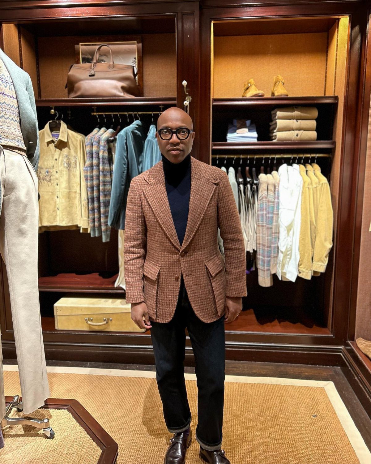 A man posing in a brown patterned blazer, navy turtleneck, and dark trousers inside a clothing store.