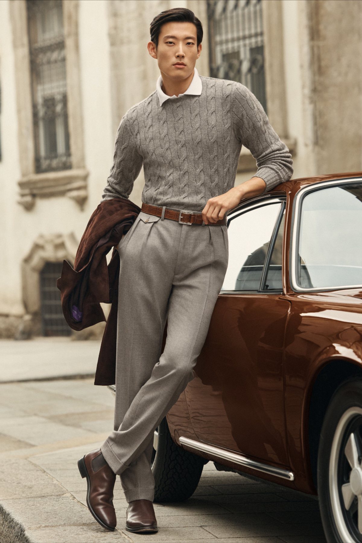 Man in a grey cable-knit sweater and grey trousers leaning against a vintage brown car.