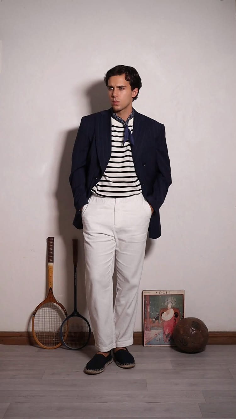 A man wearing a navy blazer, striped shirt, and white trousers standing near tennis rackets.