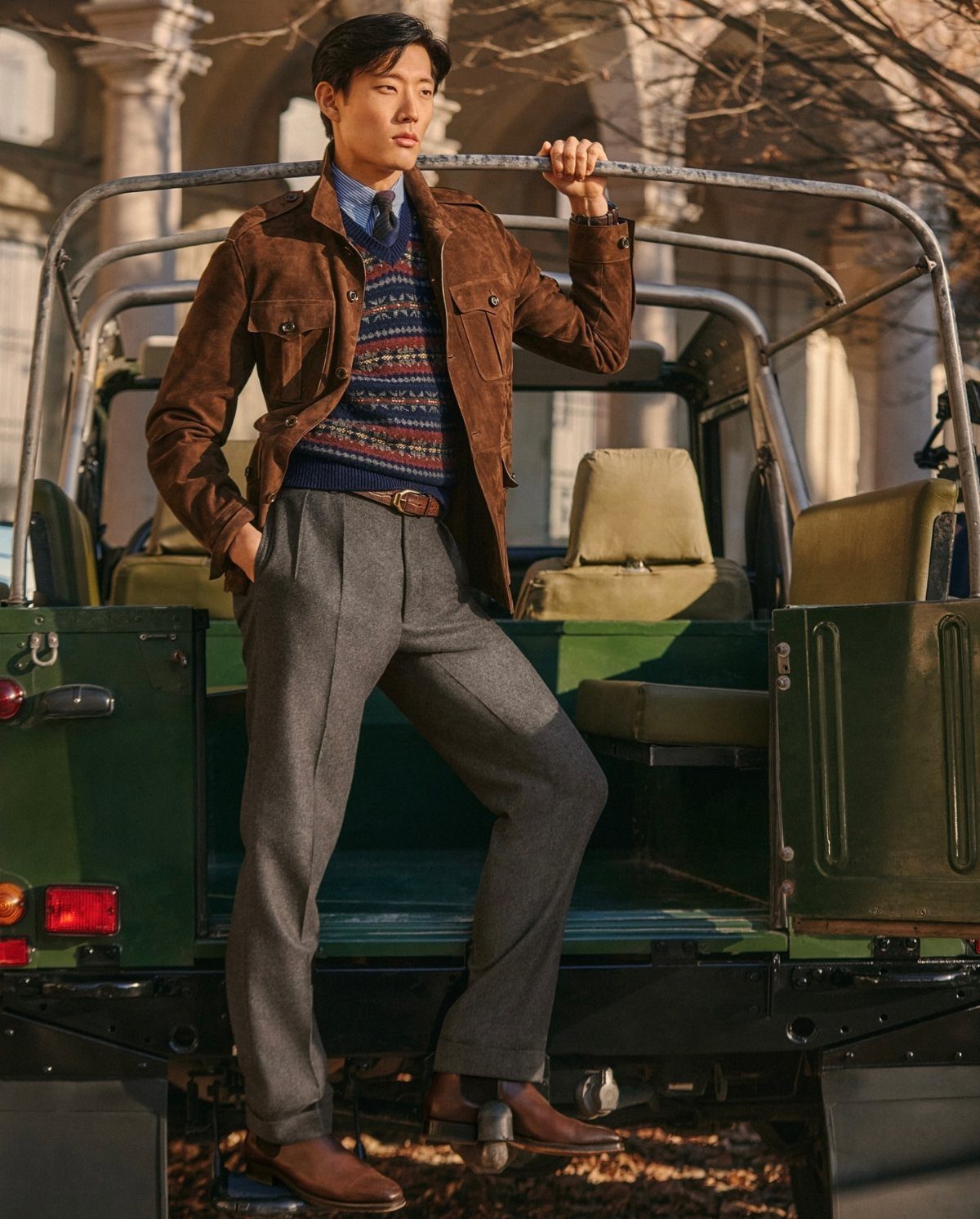 Man in a brown suede jacket and patterned sweater posing by a green vehicle.