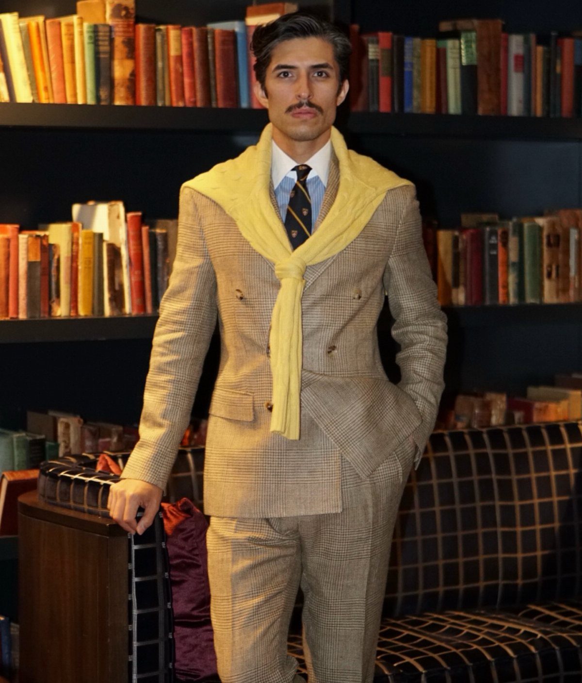 Man in a tan patterned suit with a yellow sweater draped over his shoulders standing by bookshelves.