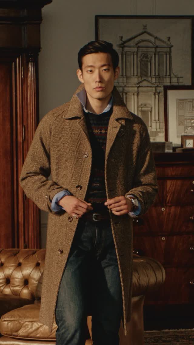 A man posing in a long brown tweed coat, patterned sweater, and dark denim jeans.