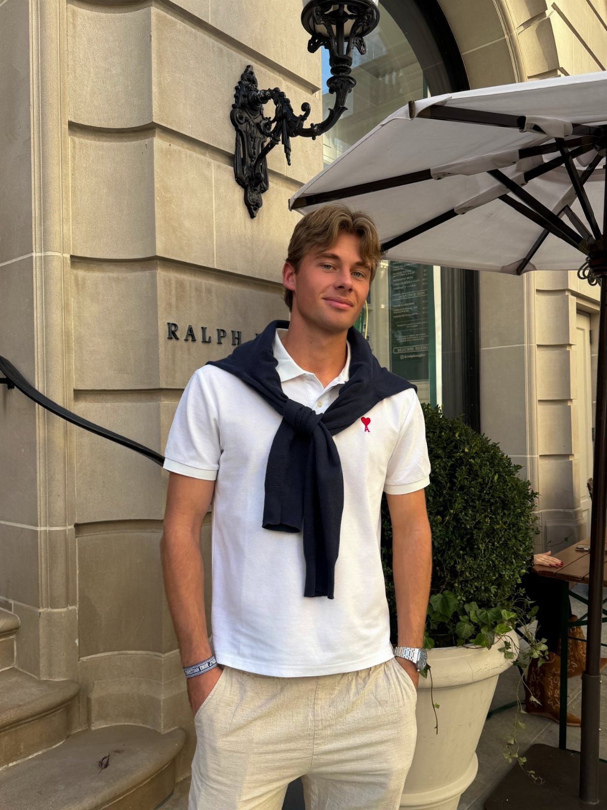 Man in white polo shirt, navy sweater over shoulders, and beige linen trousers standing outdoors.