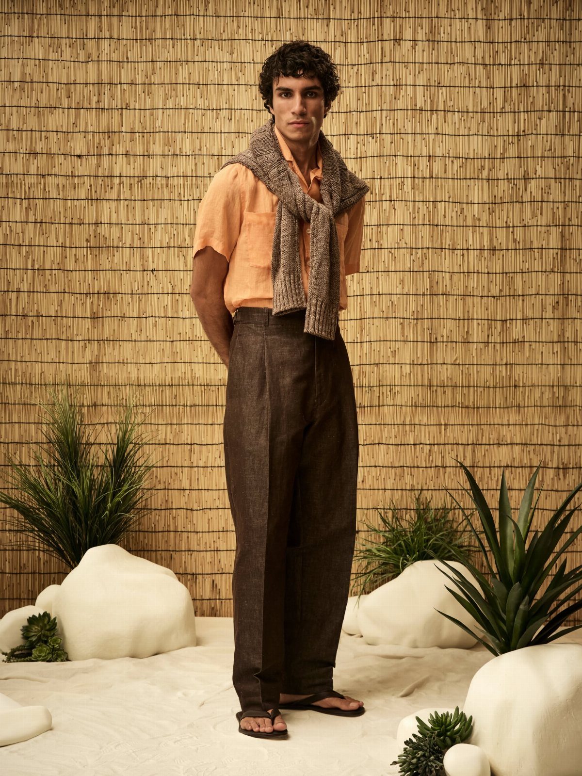 A man wearing orange shirt and dark brown relaxed trousers standing in a desert-like setting.