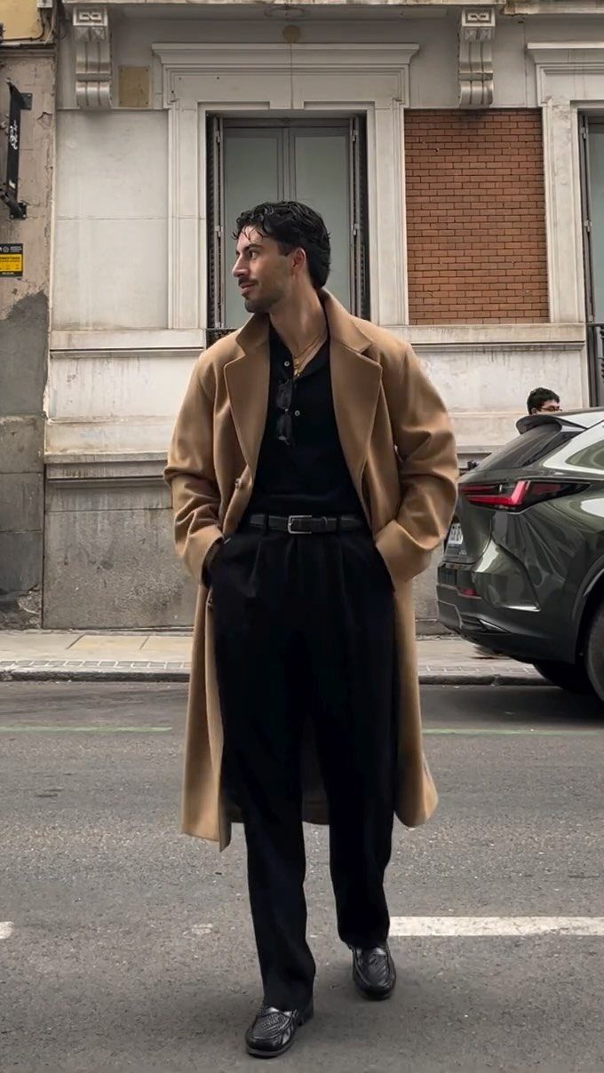 Man in a tan overcoat and black trousers standing on a city street.