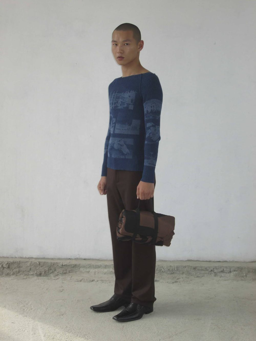 A man posing in a blue patterned knit sweater and brown trousers against a white wall.