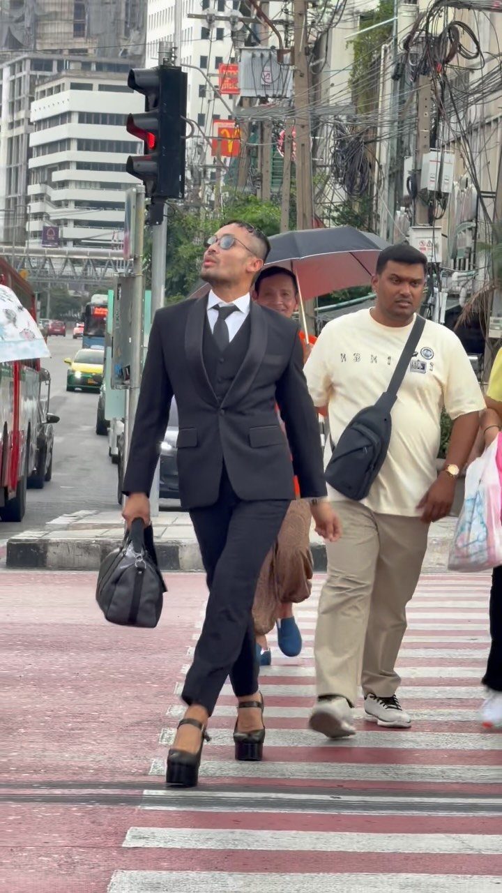 A man in a black suit walks across a street wearing very high platform sandals.