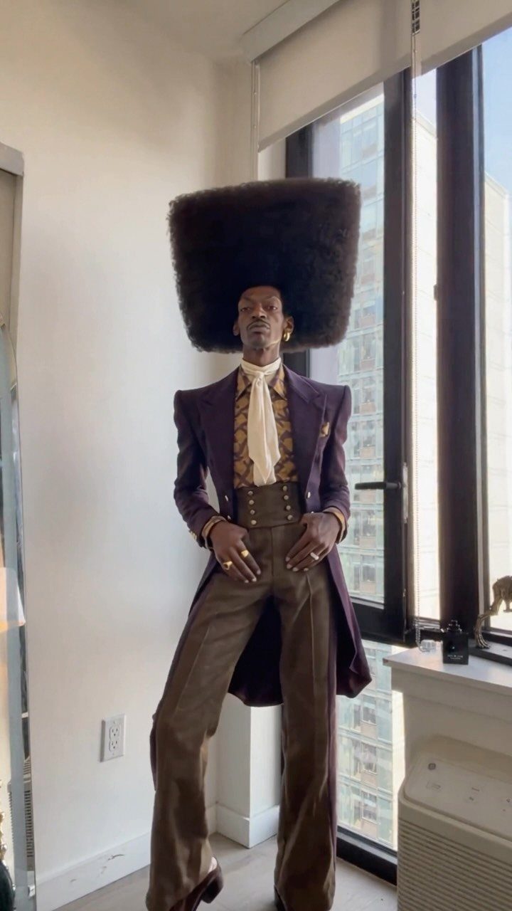 A person posing in an oversized square black fur headpiece and purple tailcoat indoors.