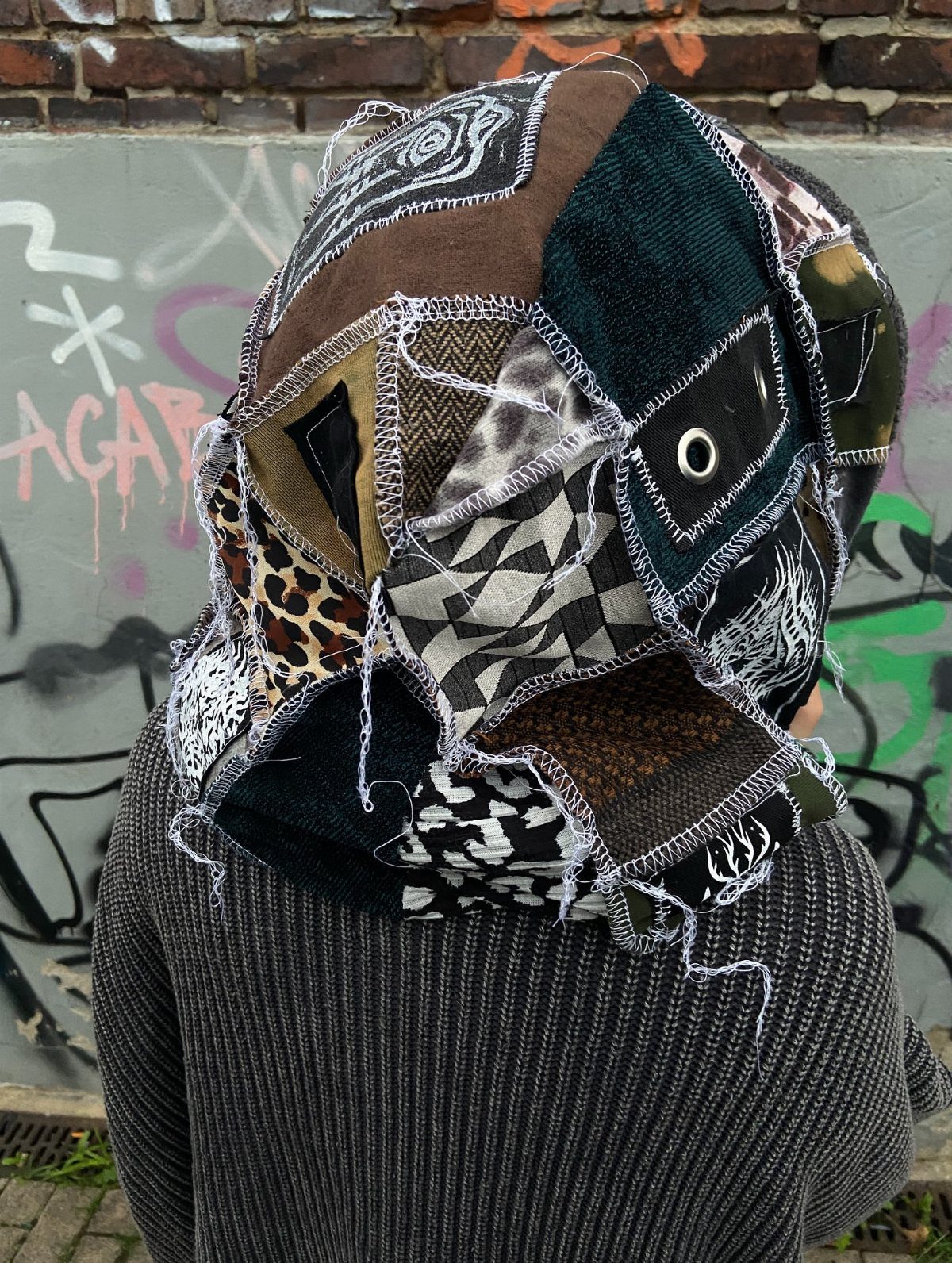 A person wearing a large, multi-patterned patchwork fabric headpiece against a graffiti wall.