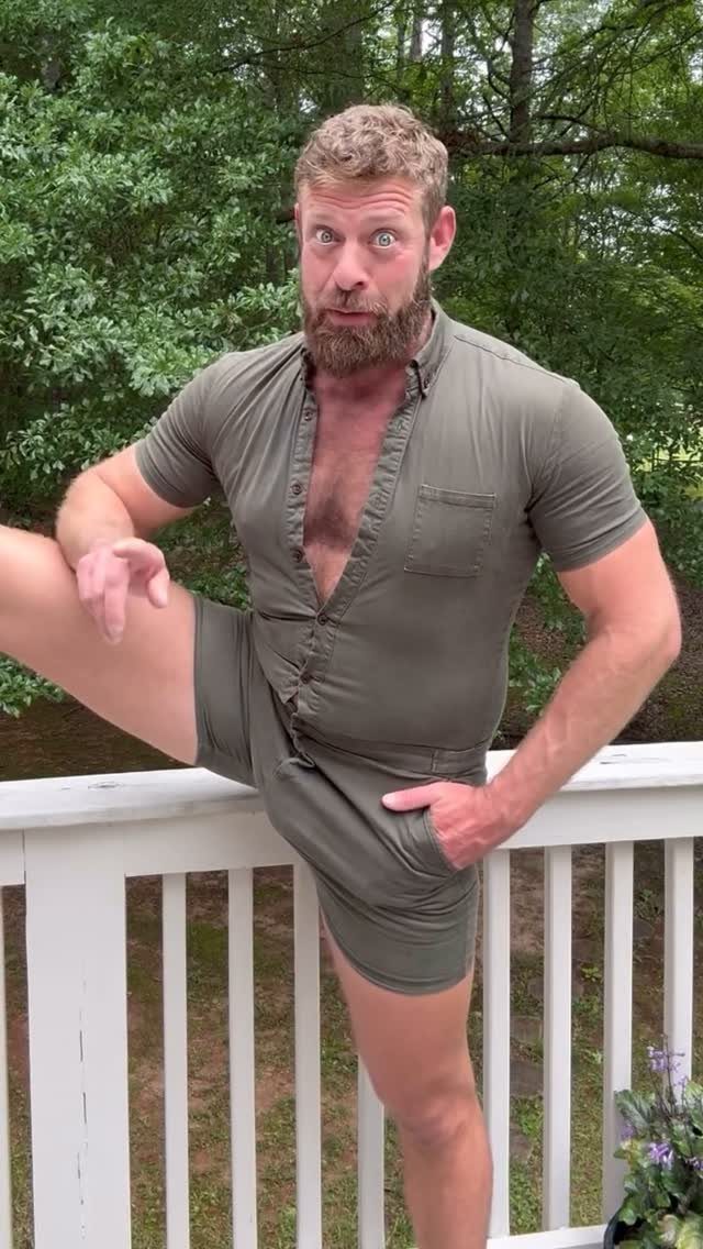 A bearded man wearing an olive green short-sleeved romper posing on a white wooden deck.