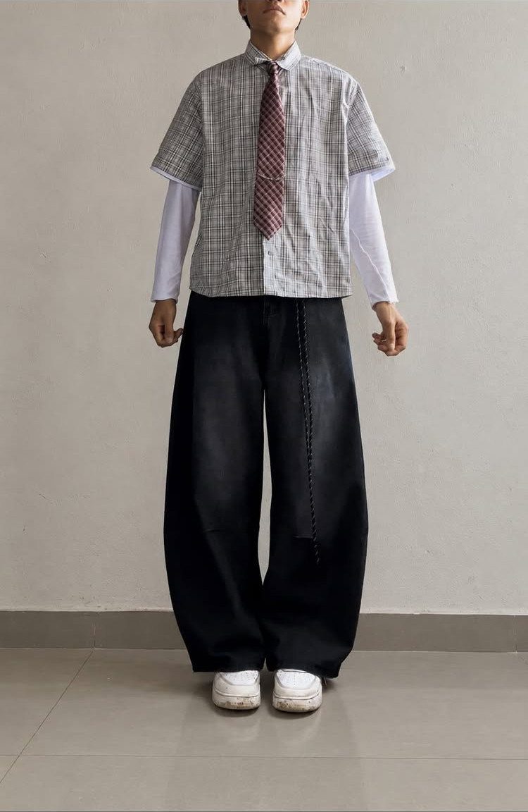 A person wearing an oversized patterned shirt, red tie, and wide black trousers against a wall.