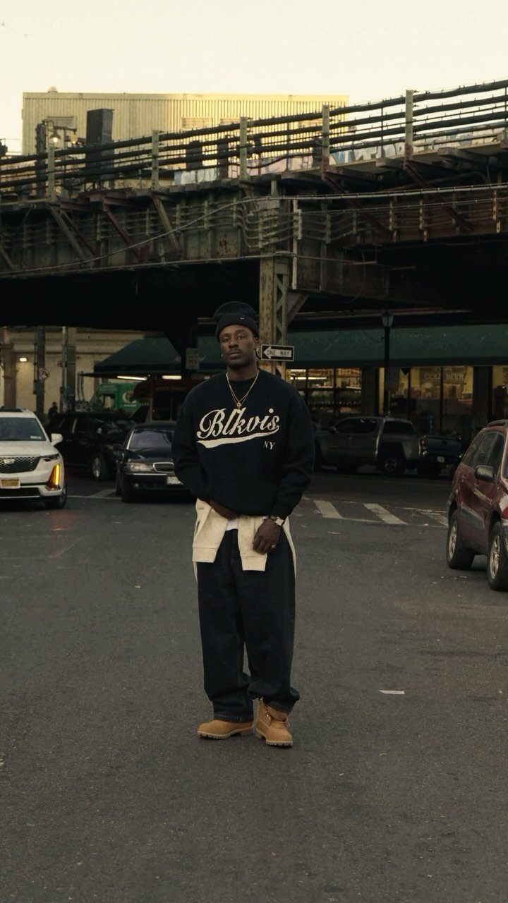 A man in streetwear standing on a city street under an elevated train track structure.