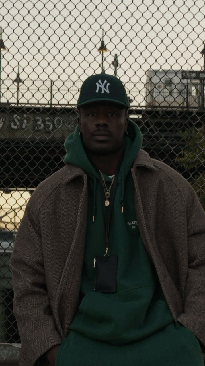 Man in a green hoodie and brown jacket standing before a chain-link fence in an urban setting.