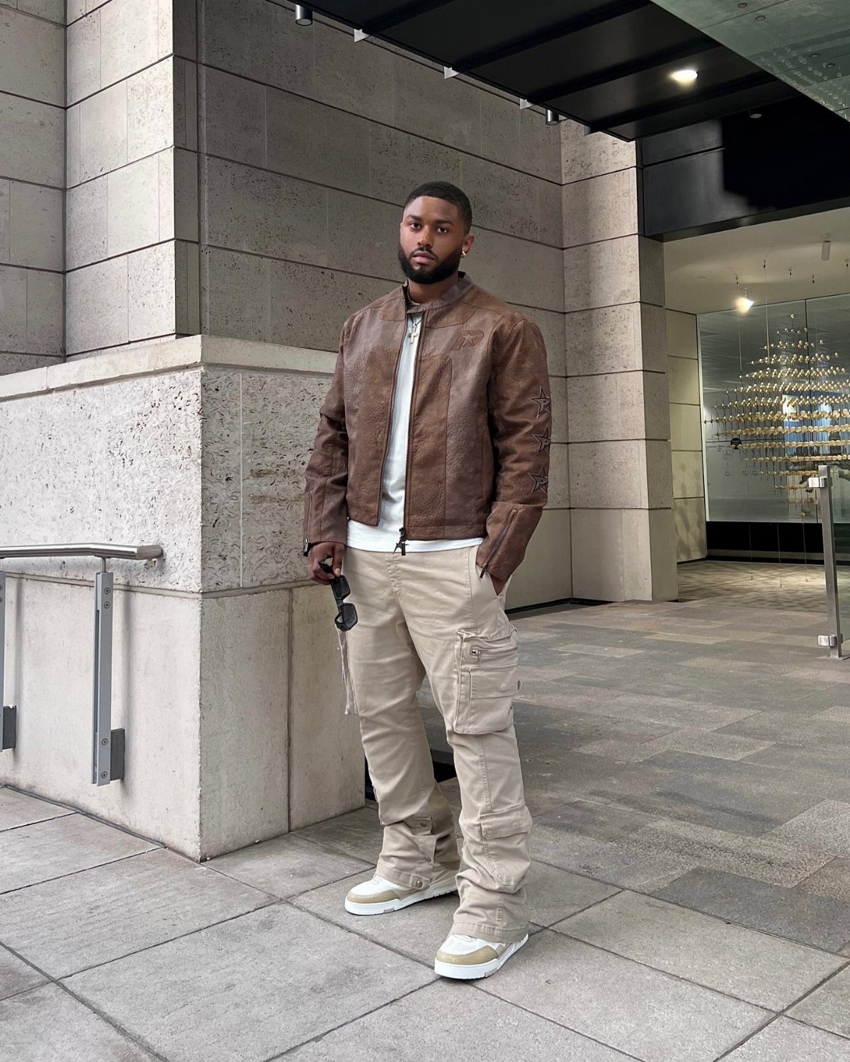 A man in a brown leather jacket and beige cargo pants posing outdoors.