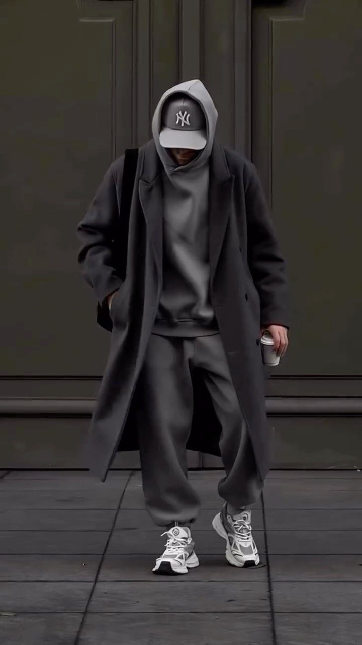 A person in monochromatic grey streetwear and a Yankees cap walking past a dark door.