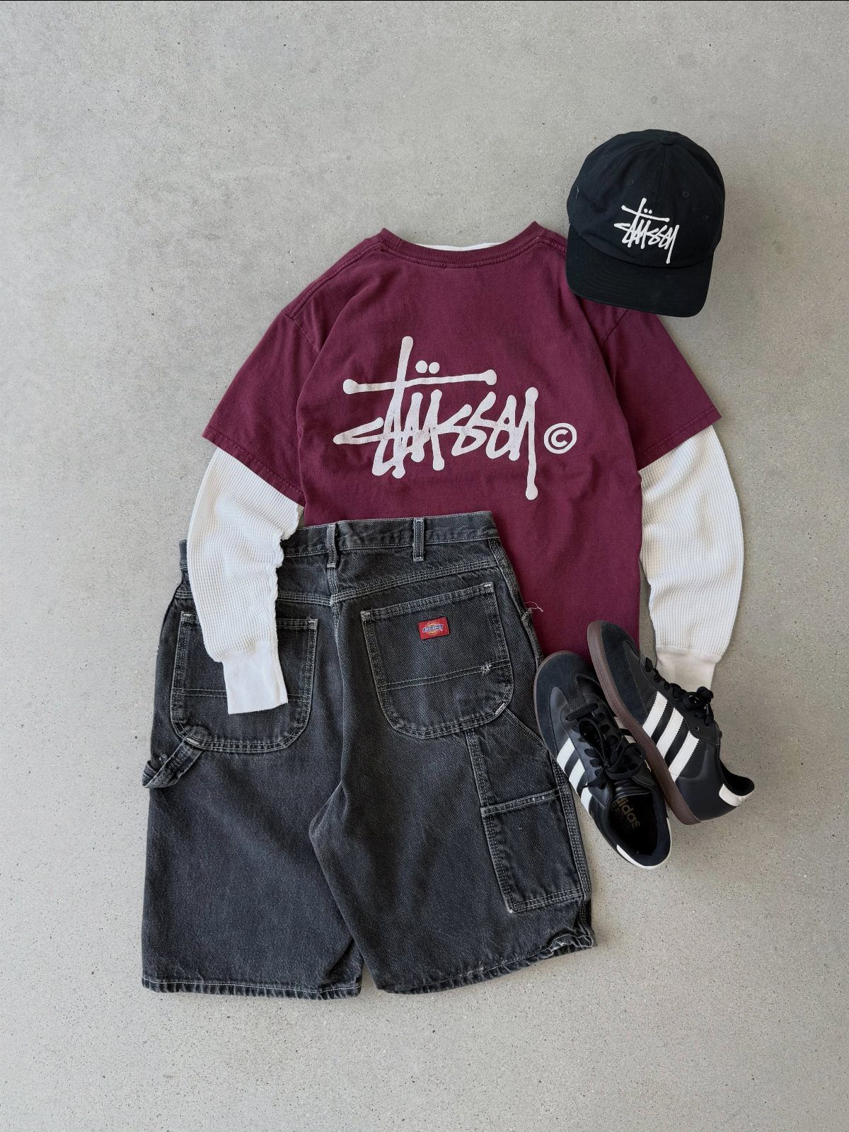 Flat lay of maroon Stüssy t-shirt, black shorts, white long-sleeve, black cap, and Adidas sneakers.
