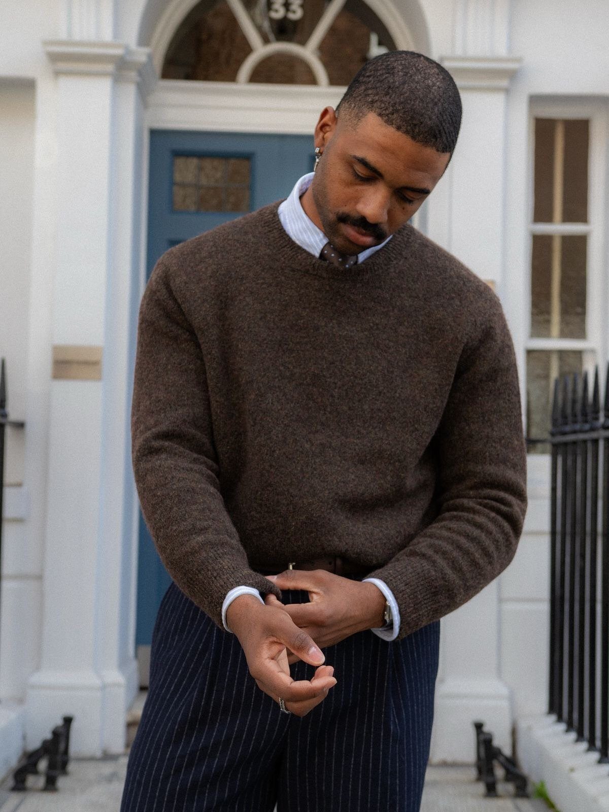 A man wearing a brown sweater and pinstriped trousers standing in front of a blue door.