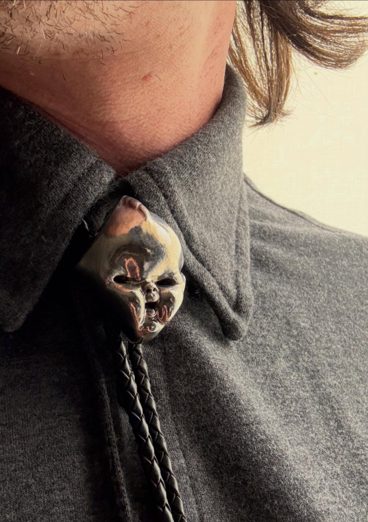A silver sculpted face bolo tie clasp on a black braided cord over a grey shirt.