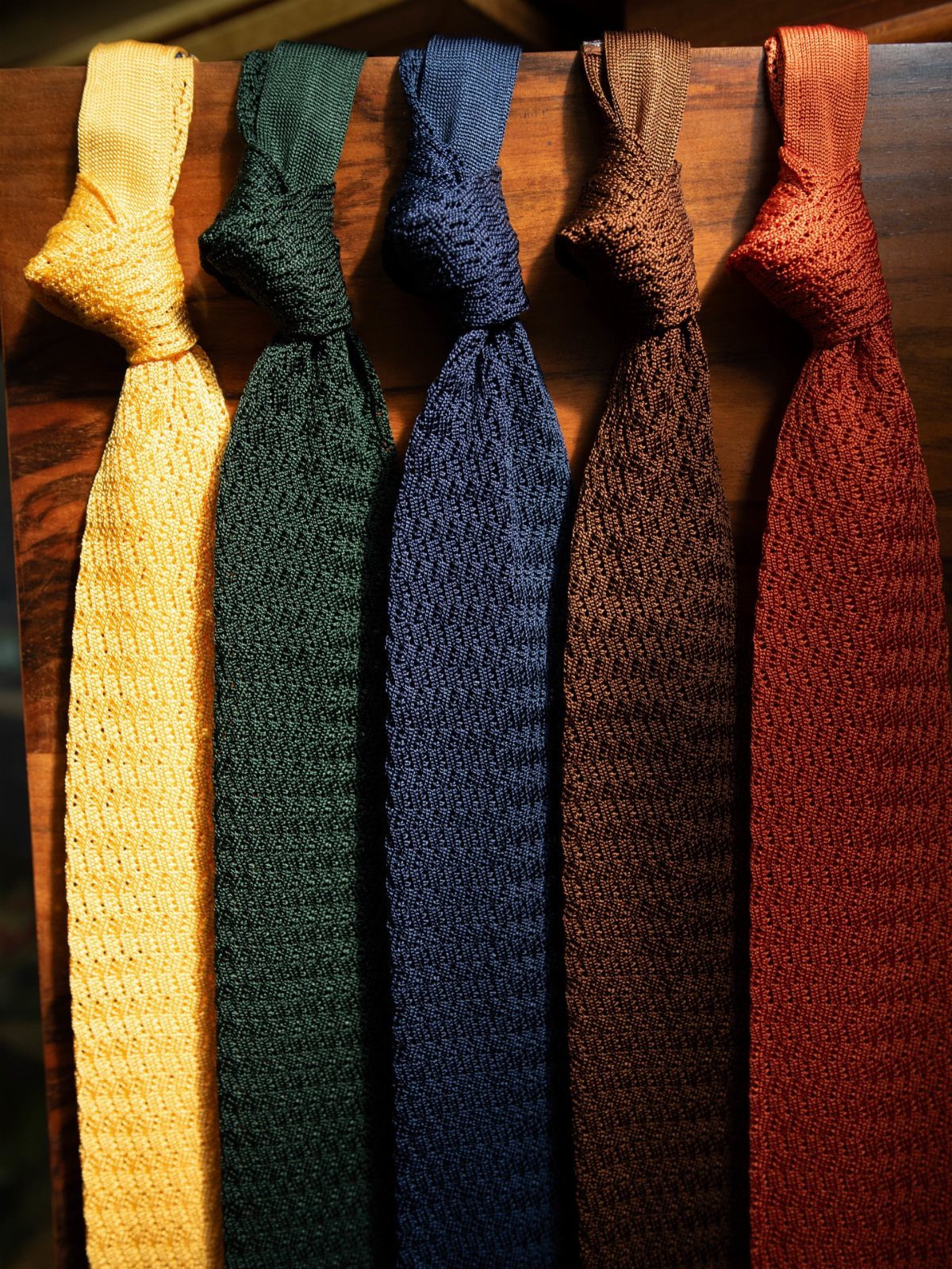 Five colorful textured neckties hanging vertically against a dark wooden surface.