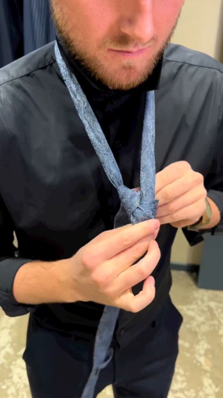 A man adjusting a light blue textured necktie while wearing a black button-down shirt.