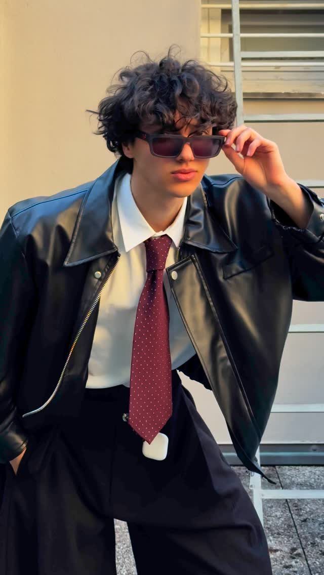 A young man with curly hair wearing a black leather jacket and a burgundy patterned necktie.
