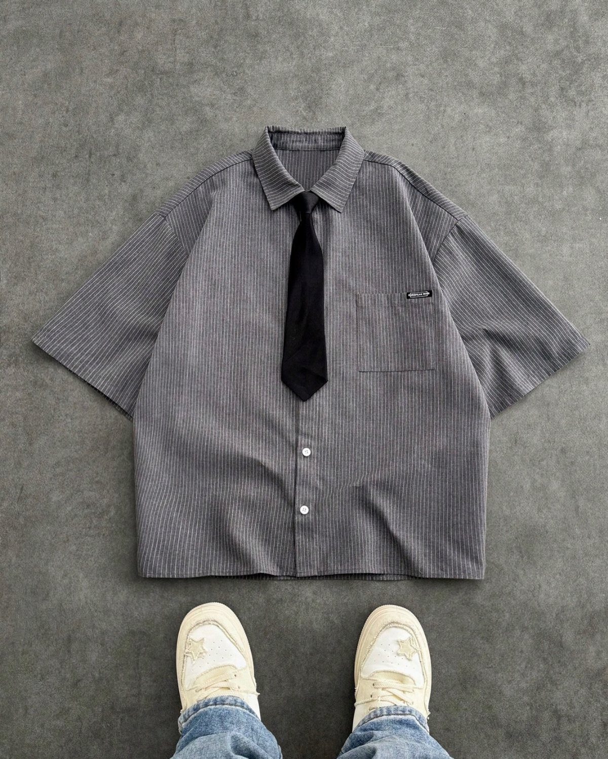 Grey striped short-sleeved shirt with a black tie laid on a grey background with sneakers.