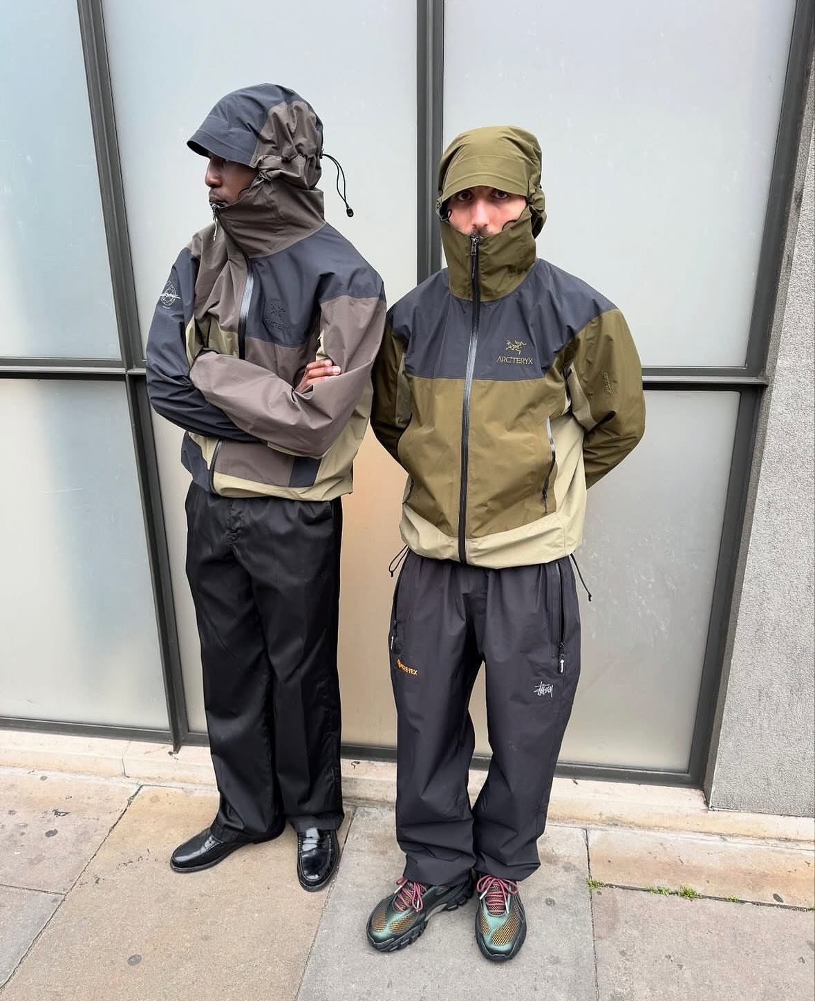 Two men posing in technical Arc'teryx jackets and outdoor trousers against a glass wall.