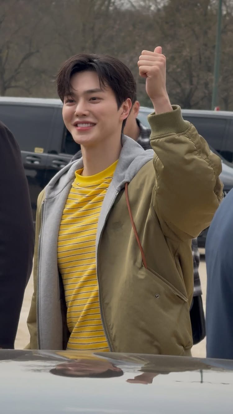 A smiling young man wearing an olive jacket, grey hoodie, and yellow striped shirt.