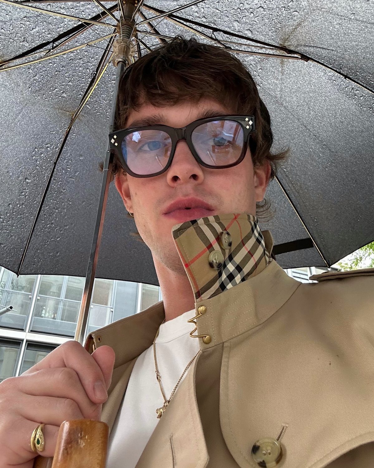 Young man wearing a tan trench coat with a plaid collar under a dark umbrella.