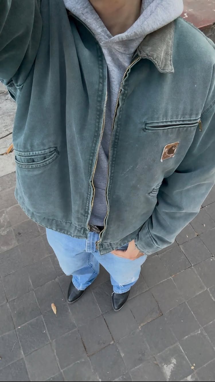 A person wearing a teal Carhartt jacket, grey hoodie, and light blue jeans standing outdoors.
