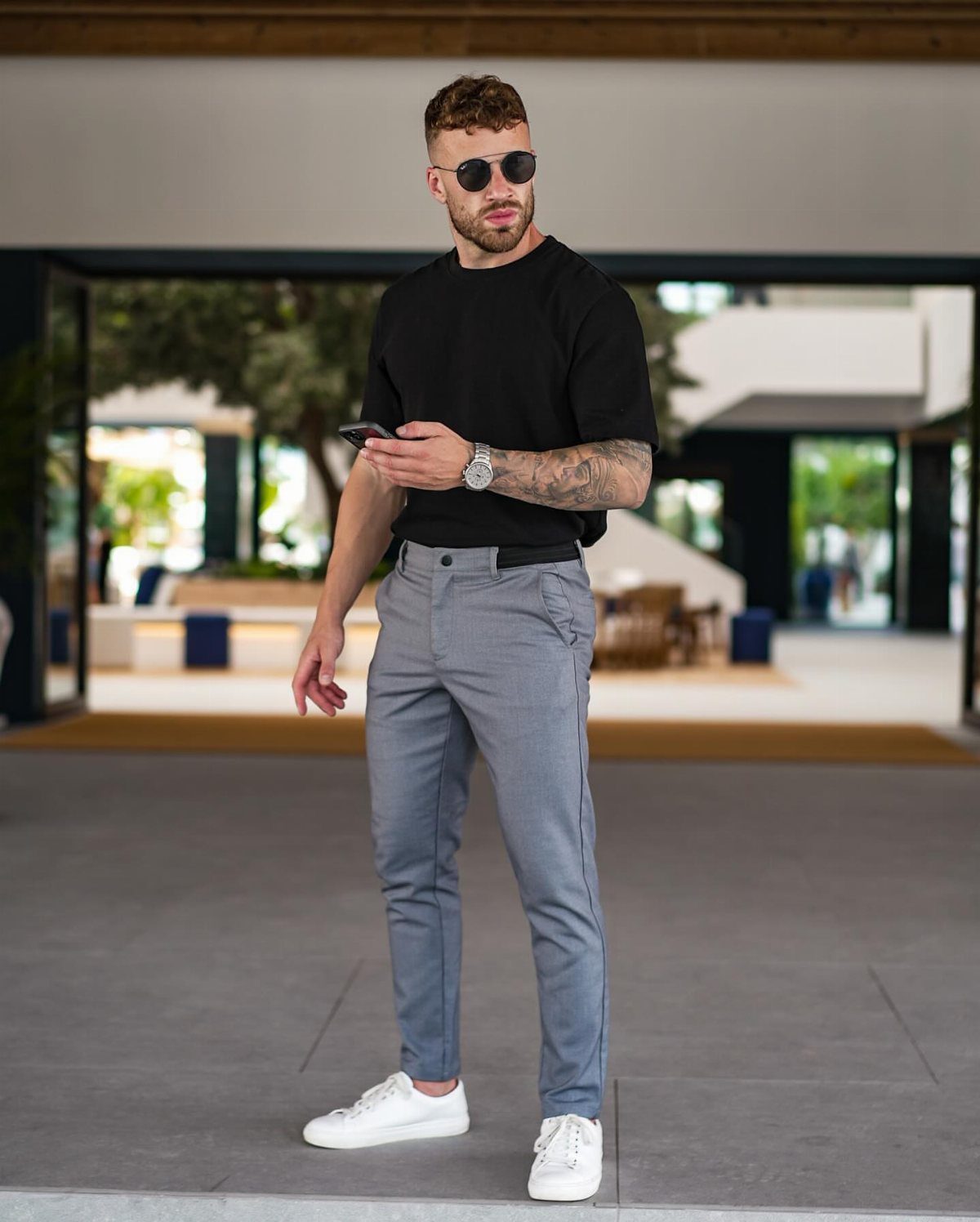 A man wearing a black t-shirt and grey chinos with white sneakers standing outdoors.