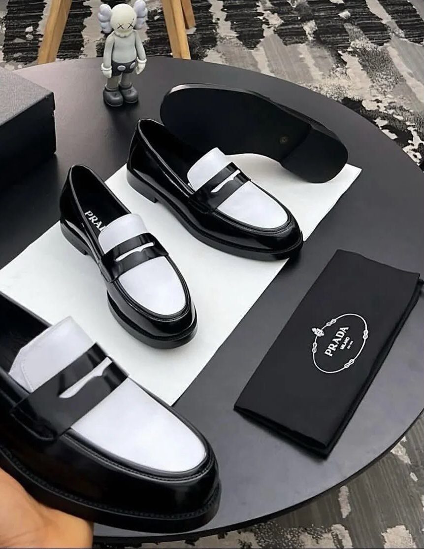 Black patent leather loafers with white inserts displayed on a dark table next to a Prada card.