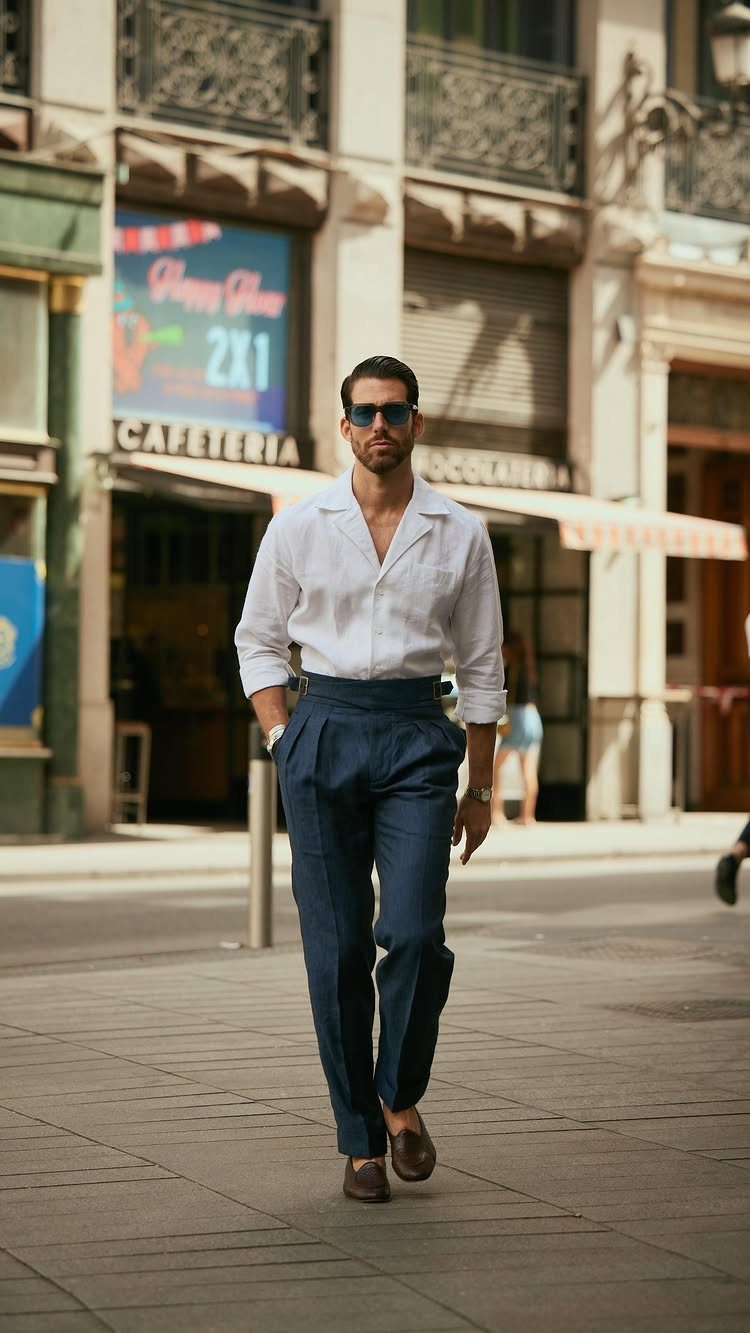 A man in a white shirt and navy trousers walking through a sunny city street.