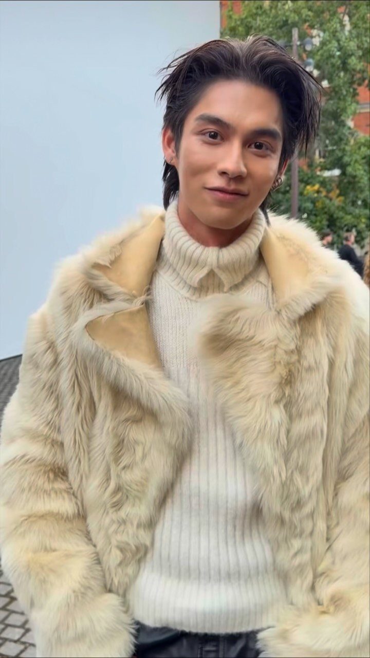 A smiling young man wearing a cream ribbed turtleneck and a large beige faux fur coat.