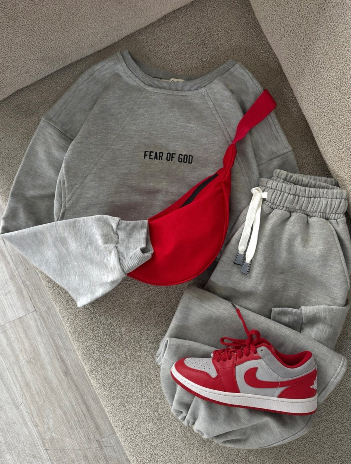 Grey Fear of God sweatshirt, sweatpants, red fanny pack, and red sneakers laid out on fabric.