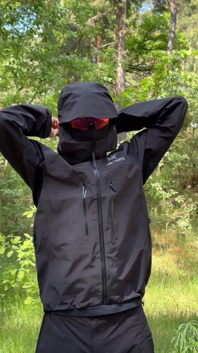 A person in black technical outdoor apparel and red sunglasses posing in a green forest.