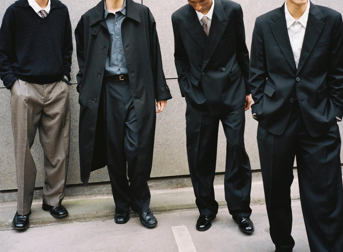 Four men standing in a row wearing minimalist, dark-toned tailored clothing and coats.
