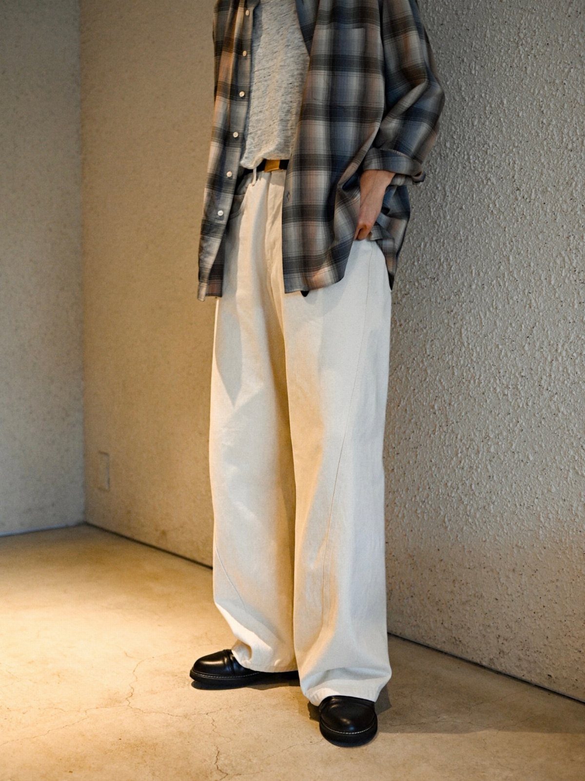 A person wearing a plaid flannel shirt, cream wide-leg trousers, and black loafers standing against a wall.