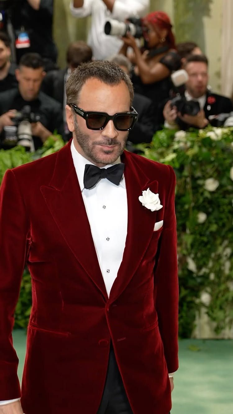 Man in a red velvet tuxedo and sunglasses walking through a crowd of photographers.