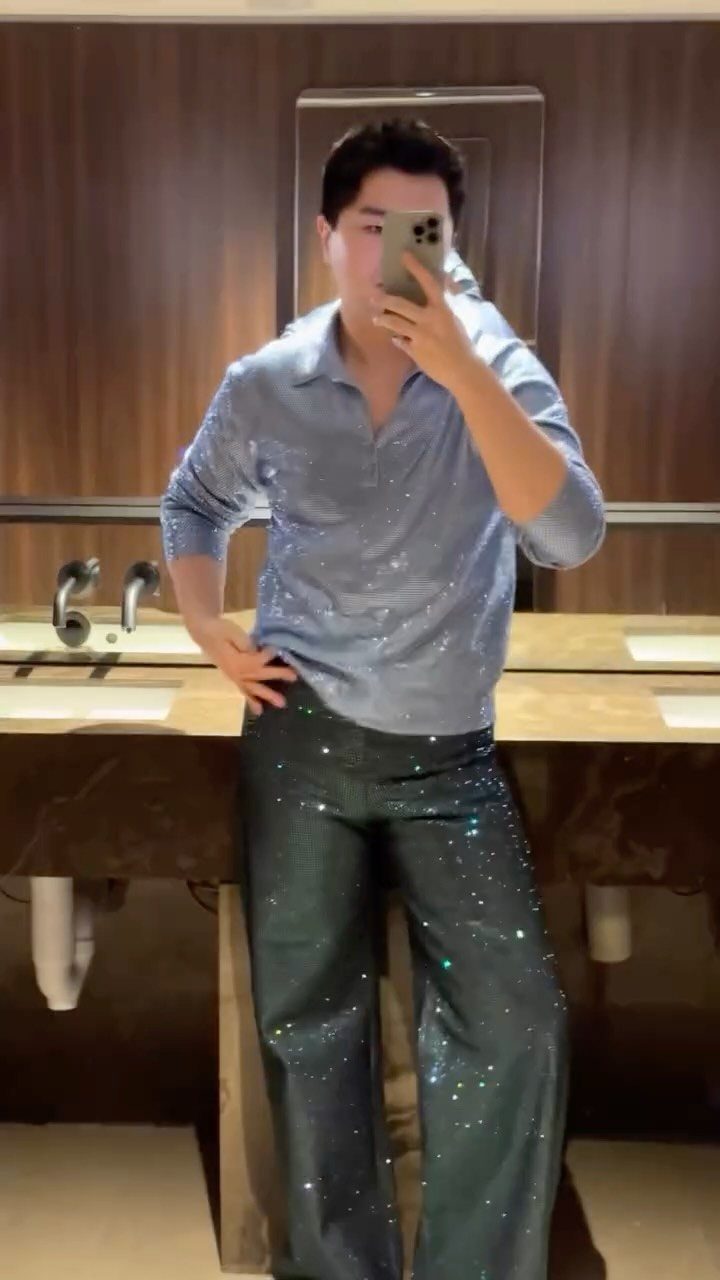 A man posing in a sequined blue shirt and glittery green trousers for a mirror selfie.