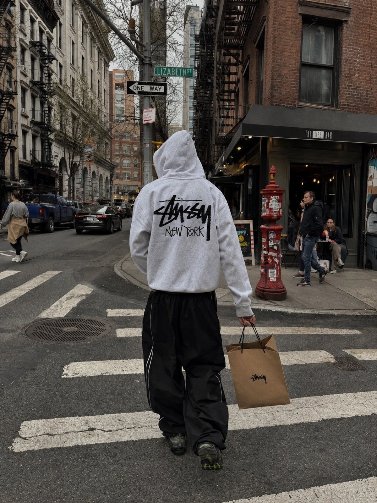 A person in a white Stüssy hoodie and black pants walking through a city street.